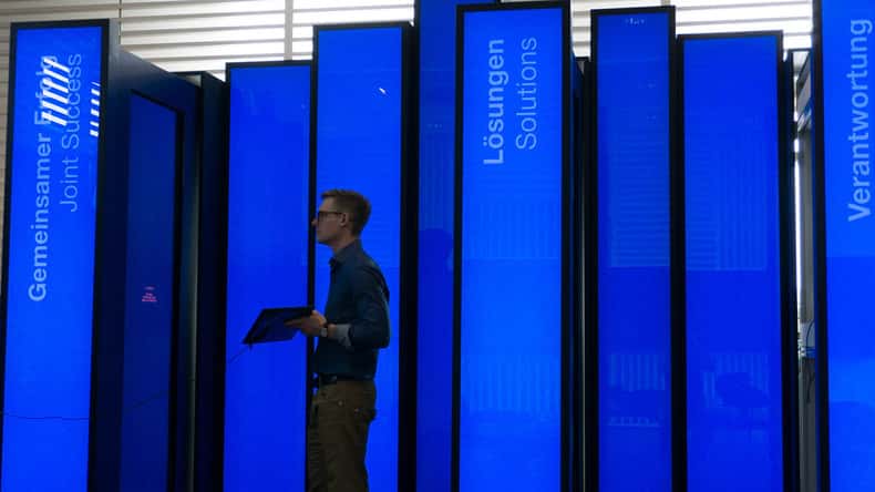 Visitor standing among tall, blue illuminated book-shaped panels of the BASF History Library, holding a tablet while navigating the spatial interactive installation inside the visitor center.