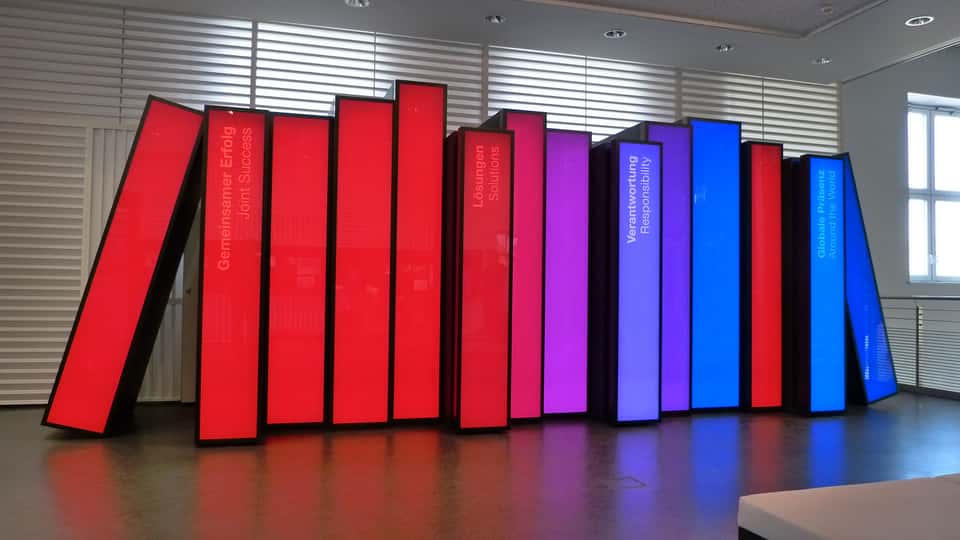 Row of illuminated, book-shaped interactive panels in red, purple, and blue forming the BASF History Library installation, representing thematic chapters of BASF’s corporate history within the visitor center.
