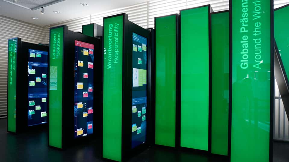 Cluster of tall, green illuminated interactive book panels displaying synchronized touchscreens, part of the BASF History Library spatial media system designed for immersive exploration of company history.