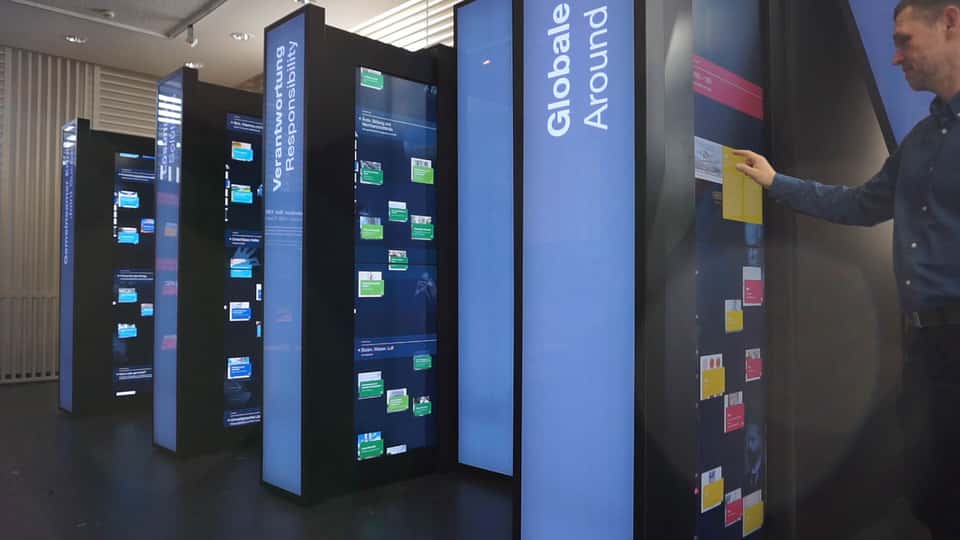 Side view of the BASF History Library installation showing multiple vertical touchscreens, with a visitor interacting with a historical content panel in a large-scale, spatially arranged media environment.