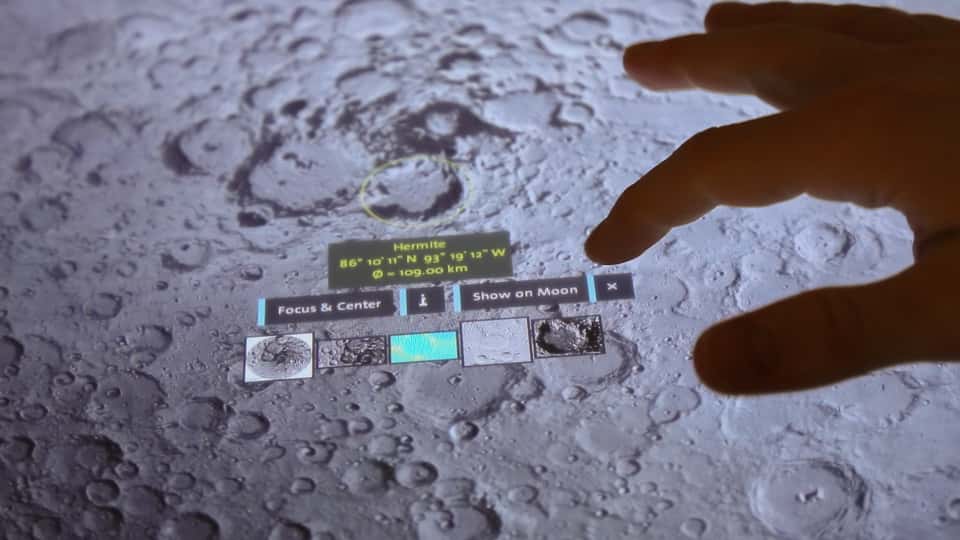 Close-up photo of a touch-enabled table displaying a 3D visualization of the moon's surface with a hand interacting with the interface.