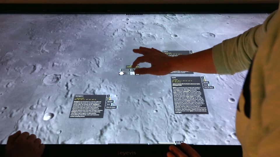 A hand doing a pinch-to-zoom gesture on the multi-touch table of the Moontable installation, zooming in on a specific area of the moon's surface displayed in 3D.