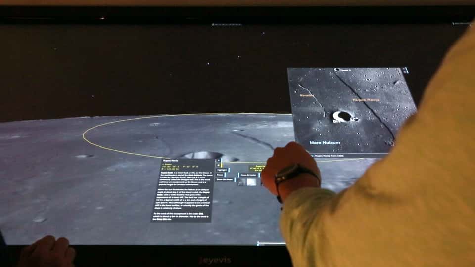 A finger pointing at a specific crater on the multi-touch table of the Moontable installation, the virtual camera being tilted to show a flat view of the moons surface.
